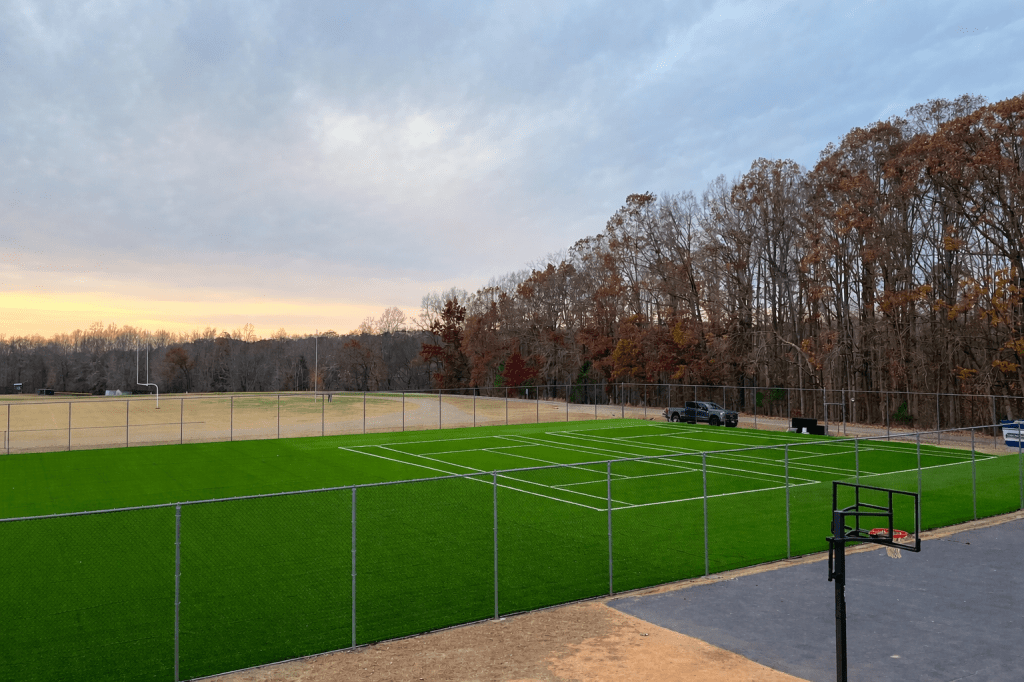 An outdoor sports field with a basketball hoop and a football field, surrounded by trees.