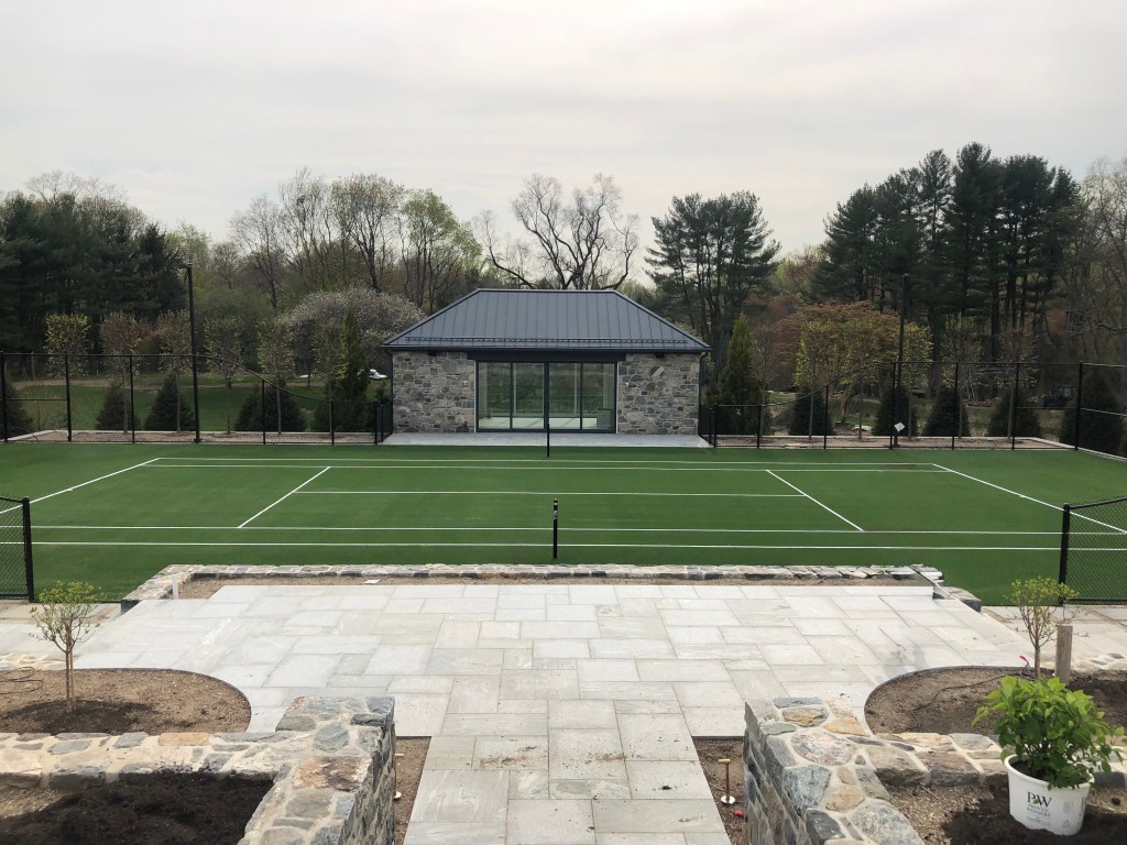 The image depicts a tennis court with a small stone building in the background, surrounded by trees and a cloudy sky. The ...